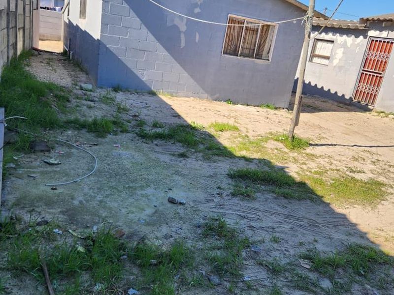 Rear exterior view of a concrete block house featuring a security gate and perimeter wall, showing signs of wear and unmaintained grounds.