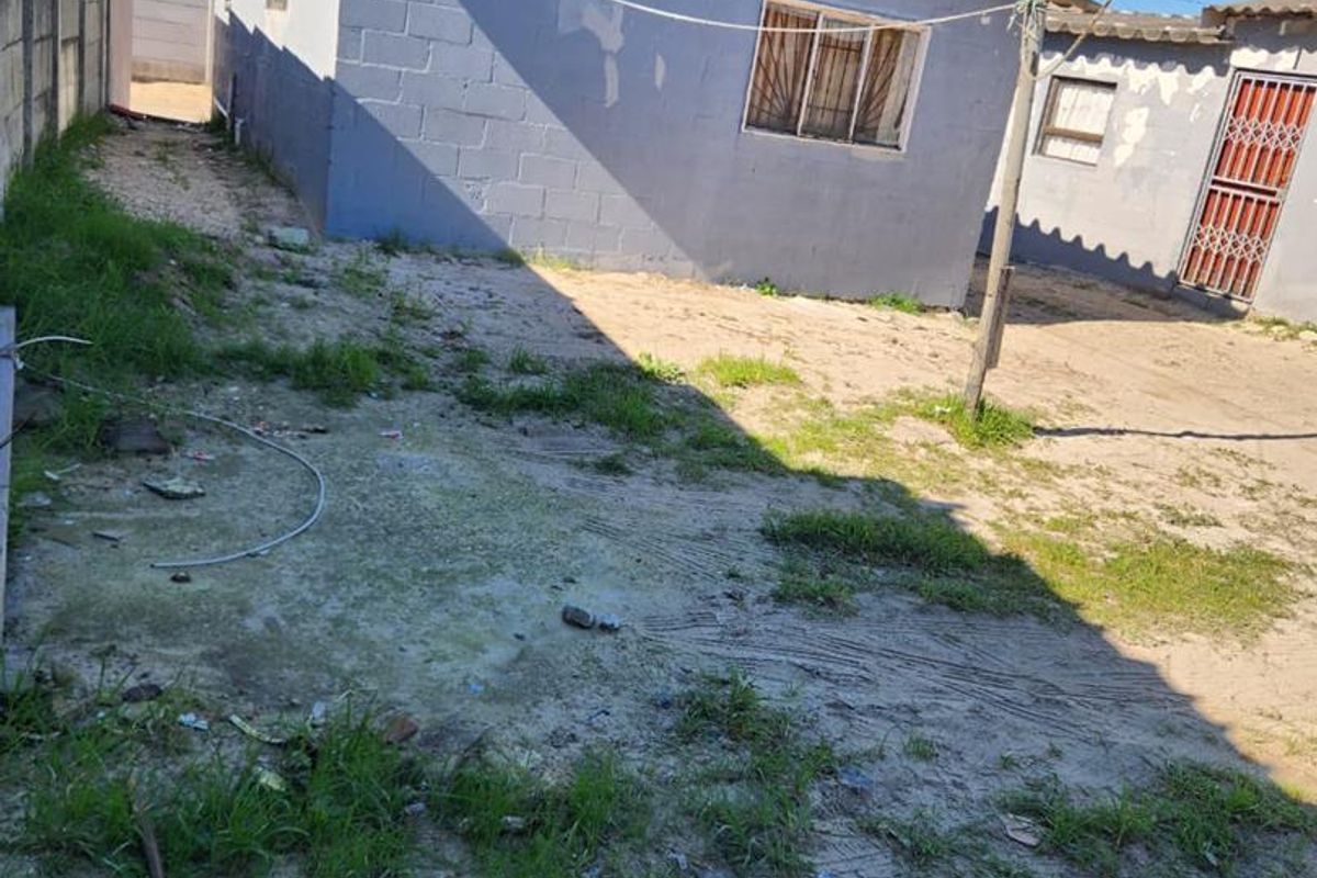 Rear exterior view of a concrete block house featuring a security gate and perimeter wall, showing signs of wear and unmaintained grounds.