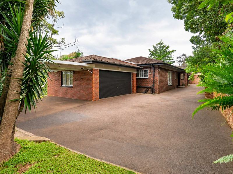 Front exterior view of a brick townhouse featuring a double garage, attached carport, and a spacious paved driveway surrounded by established garden greenery.