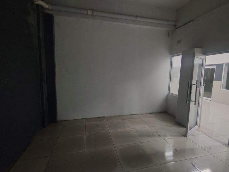 An empty, utilitarian office space featuring tiled flooring, exposed ceiling pipes, and an aluminium-framed glass entrance door.