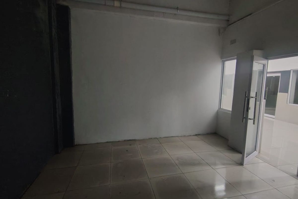 An empty, utilitarian office space featuring tiled flooring, exposed ceiling pipes, and an aluminium-framed glass entrance door.