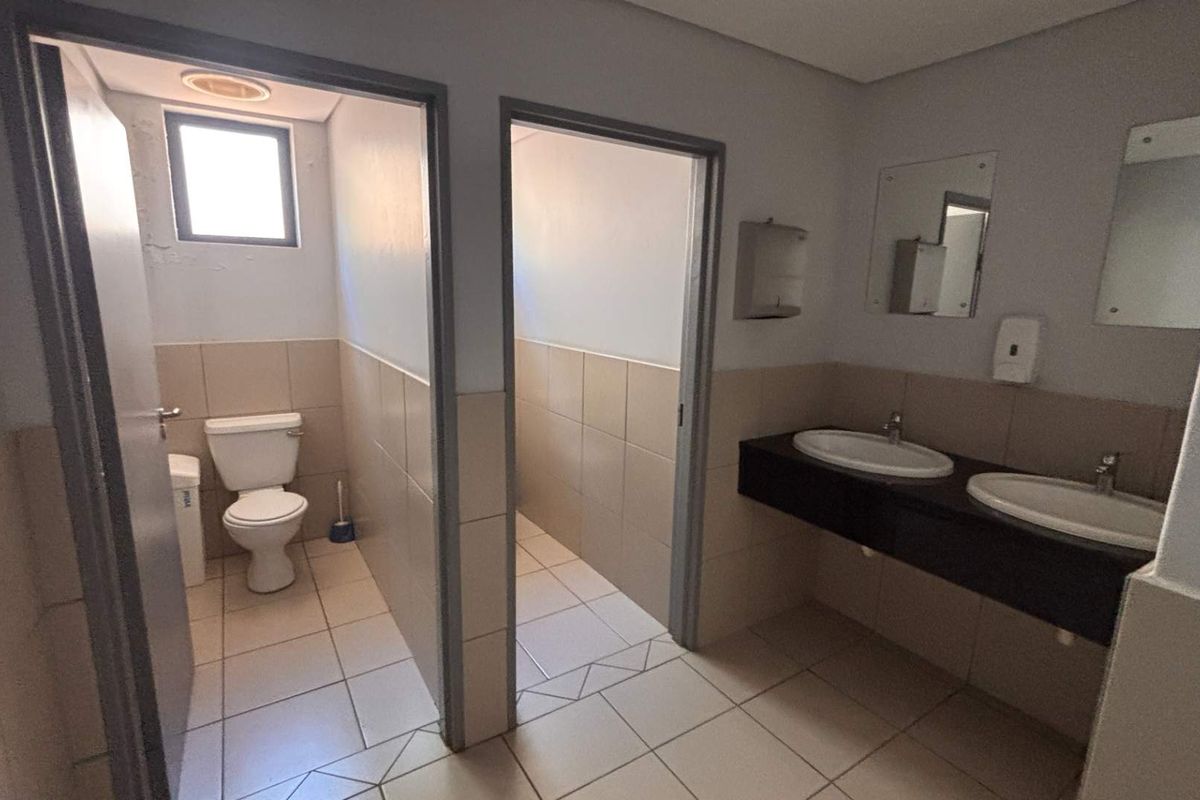 Functional office bathroom featuring a dual vanity with integrated sinks, tiled walls and flooring, and a separate enclosed toilet cubicle.
