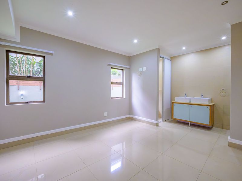 A spacious, well-lit bedroom featuring neutral tiled flooring, recessed downlights, and an integrated double vanity area with built-in cabinetry.