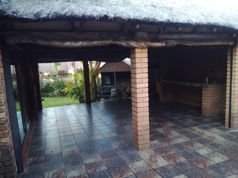 Covered outdoor patio area featuring a thatched roof, brick support pillars, and a built-in brick braai, overlooking the garden.