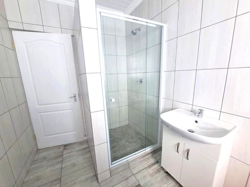 A clean, modern bathroom featuring a glass-enclosed shower, a white vanity cabinet with a basin, and neutral floor-to-ceiling wall tiles.