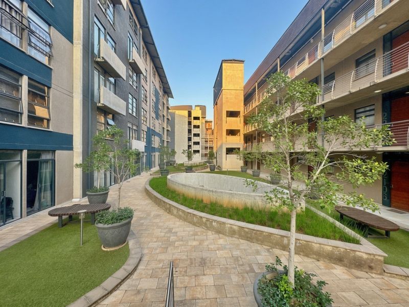 A well-maintained central courtyard area between apartment blocks featuring paved walkways, landscaped garden beds, and modern building facades with balconies.