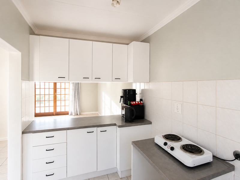 A functional kitchen featuring white cabinetry, grey countertops, tiled backsplash, and a compact electric hob, with a view through to the adjacent living area.