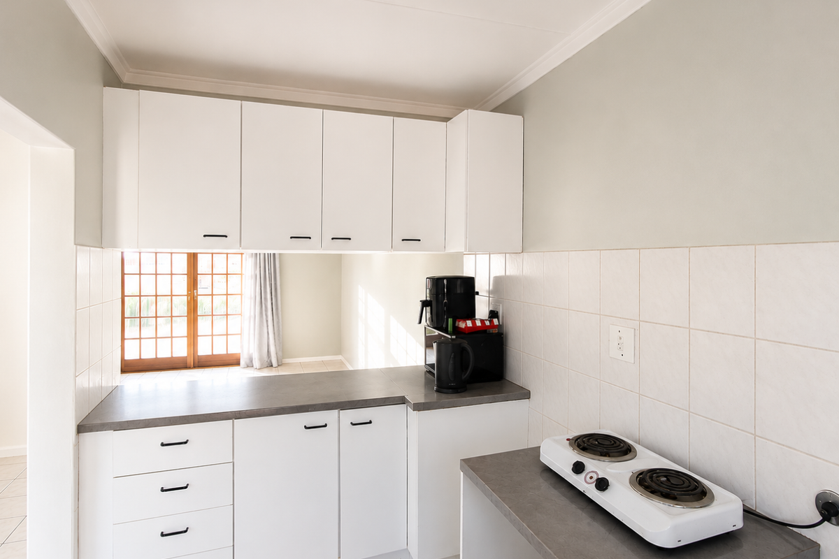 A functional kitchen featuring white cabinetry, grey countertops, tiled backsplash, and a compact electric hob, with a view through to the adjacent living area.