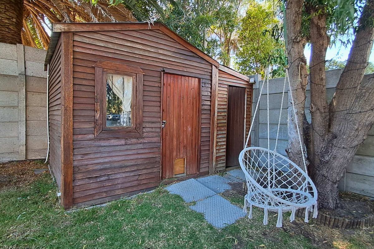 A detached wooden garden shed or utility structure situated in a backyard setting, featuring timber cladding and a concrete boundary wall backdrop.
