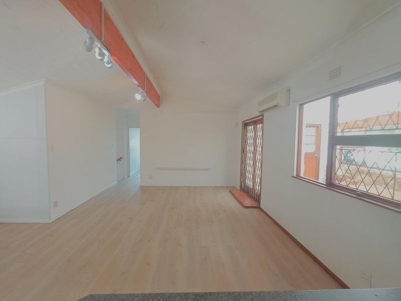 An open-plan living area featuring light laminate flooring, an exposed timber beam, and a wall-mounted air conditioning unit with access to an outdoor space.