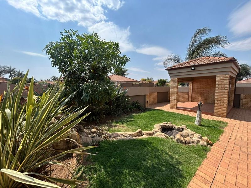 A well-maintained rear garden featuring a covered brick patio, paved walkways, and a stone-lined fire pit area within a secure cluster property.
