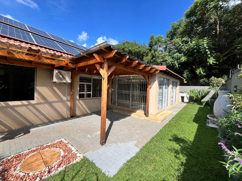A well-maintained paved patio area featuring a wooden pergola, security gates, and access to a landscaped garden with solar panels installed on the roof.