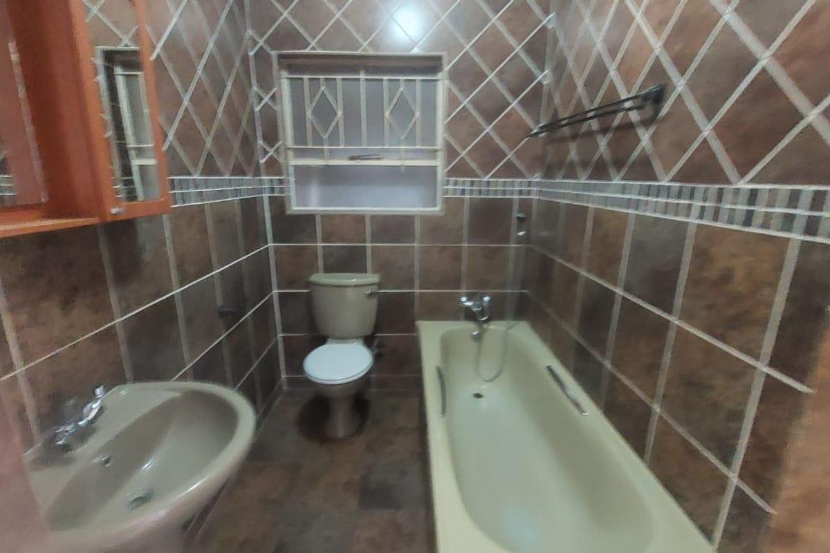A dated bathroom featuring full-height wall tiling, a standard bathtub, pedestal basin, and toilet, with security bars installed on the window.