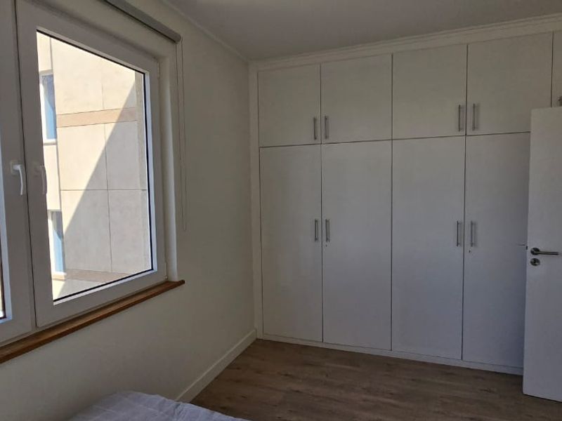 A bright, empty bedroom featuring modern white built-in cupboards, laminate flooring, and large windows offering a clear exterior view.