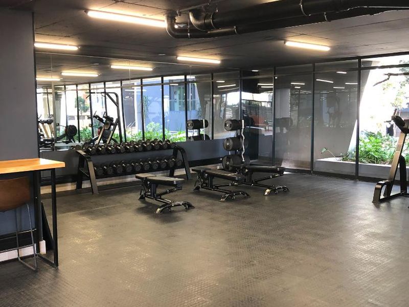 A functional apartment gym facility featuring rubber flooring, mirrored walls, and exposed industrial-style ceiling pipes with integrated lighting.