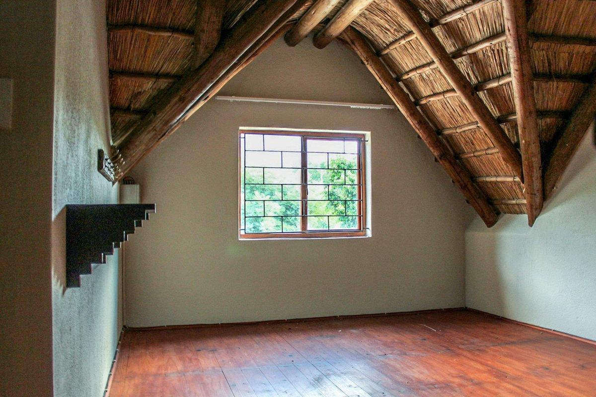 An empty attic-style bedroom featuring exposed timber rafters, a thatched roof ceiling, and a window with security bars.