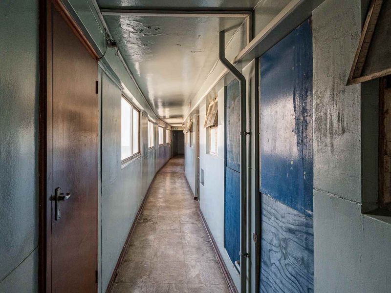 A long, narrow apartment hallway featuring tiled flooring, multiple access doors, and exposed utility piping, currently showing significant signs of wear and dated finishes.