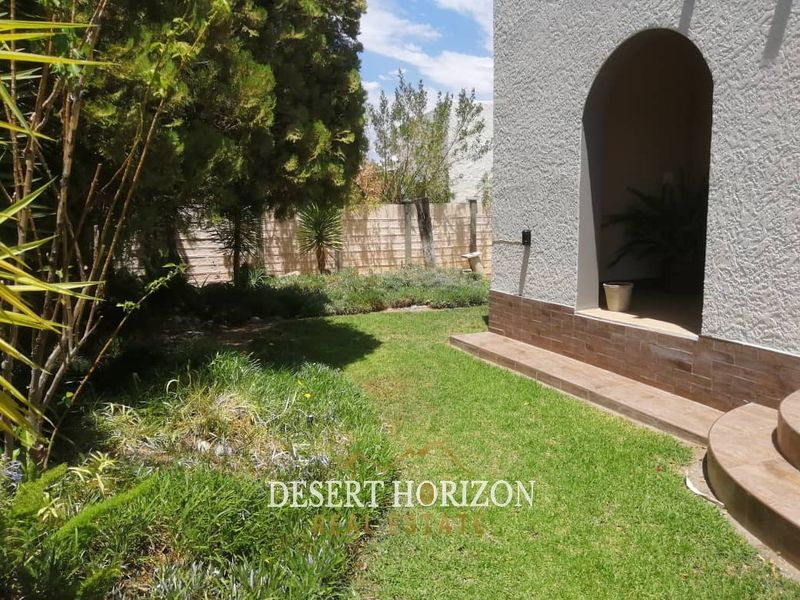 A view of the rear garden featuring a landscaped lawn, boundary wall, and a covered arched entryway with tiled steps.