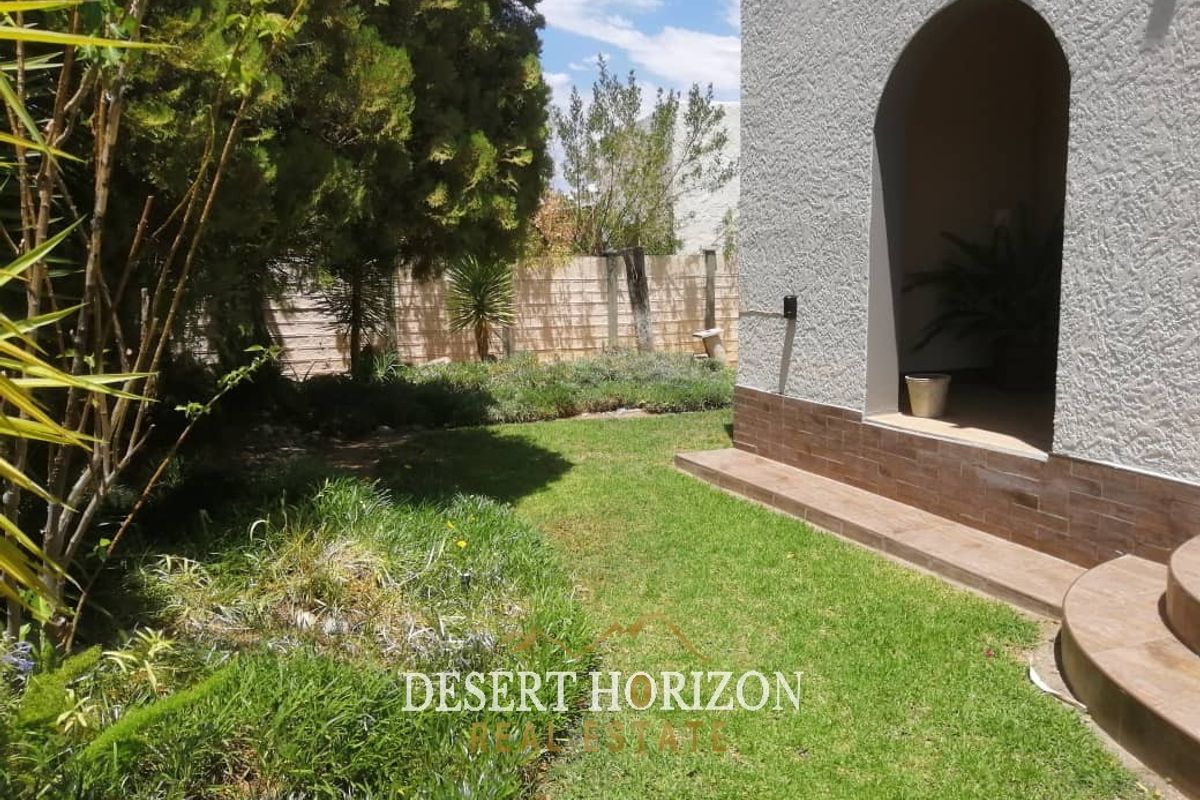 A view of the rear garden featuring a landscaped lawn, boundary wall, and a covered arched entryway with tiled steps.
