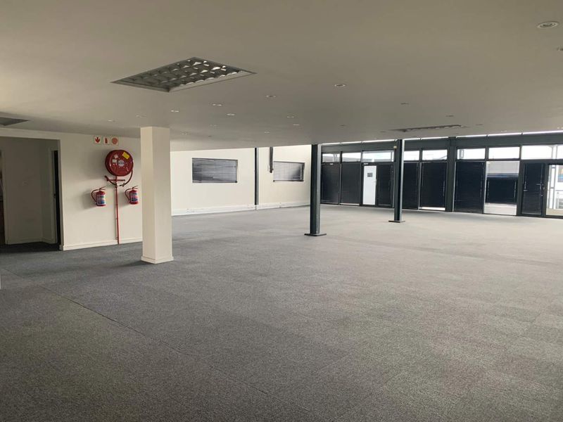 A spacious, open-plan office area featuring carpeted flooring, structural columns, recessed lighting, and large windows with fitted blinds.