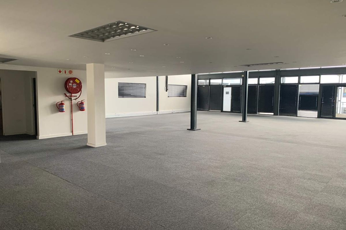 A spacious, open-plan office area featuring carpeted flooring, structural columns, recessed lighting, and large windows with fitted blinds.