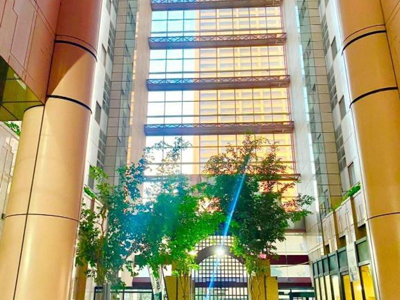 A grand, high-ceilinged building atrium featuring extensive glass paneling, structural columns, and stone flooring, creating a bright and modern entrance space.