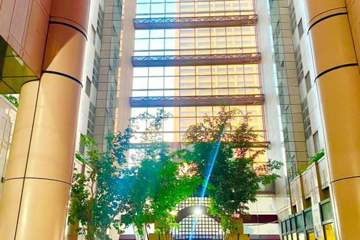 A grand, high-ceilinged building atrium featuring extensive glass paneling, structural columns, and stone flooring, creating a bright and modern entrance space.
