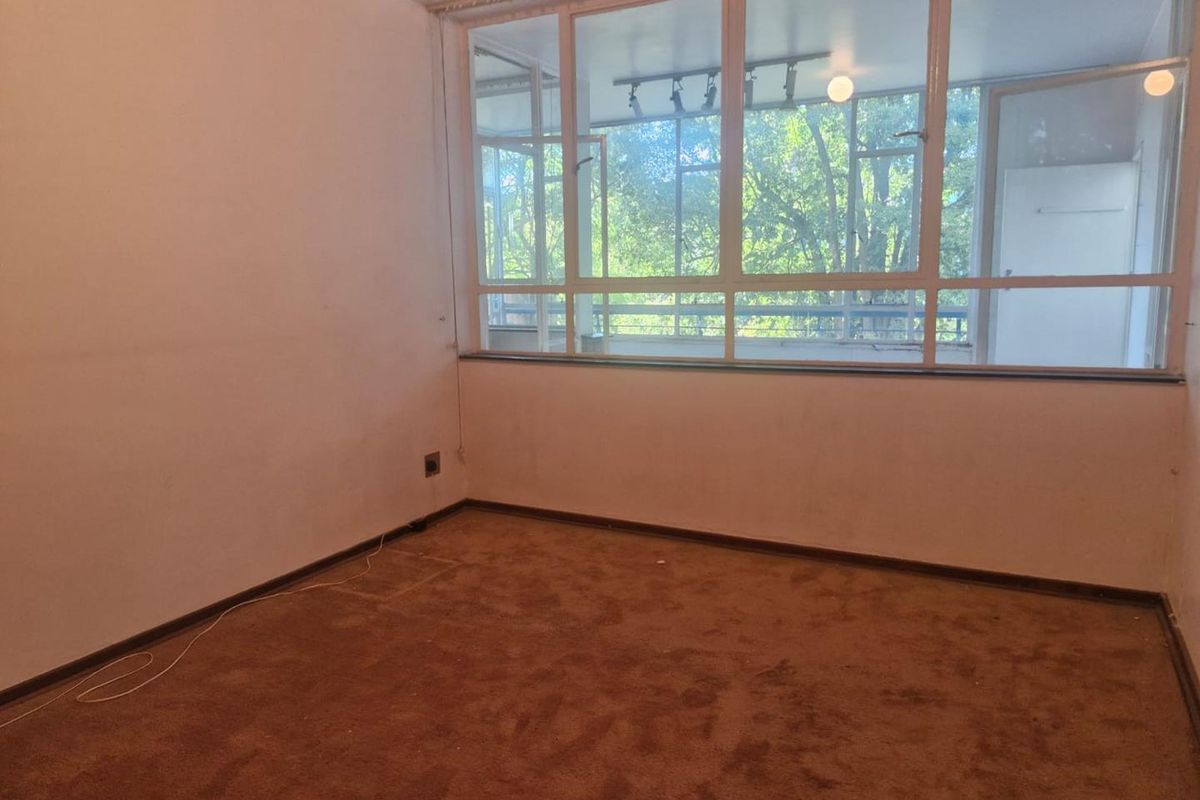 An empty bedroom featuring brown carpeting, plain walls, and large windows providing natural light and a view of the surrounding trees.