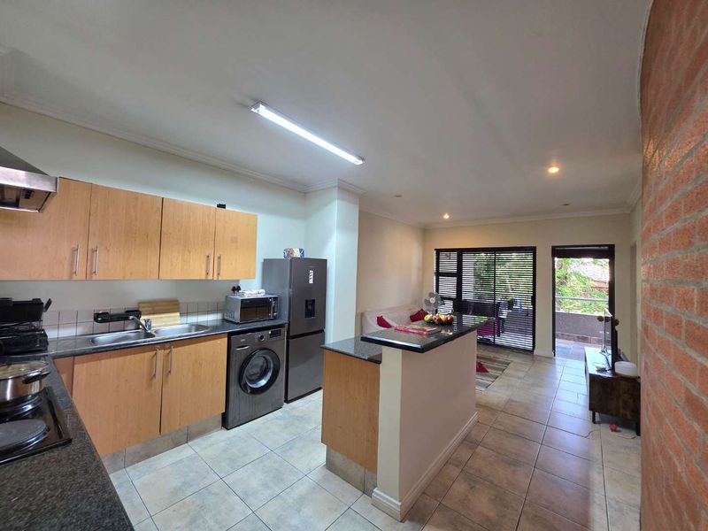 Open-plan kitchen and living area featuring light wood cabinetry, granite countertops, tiled floors, and a breakfast bar leading to a balcony.