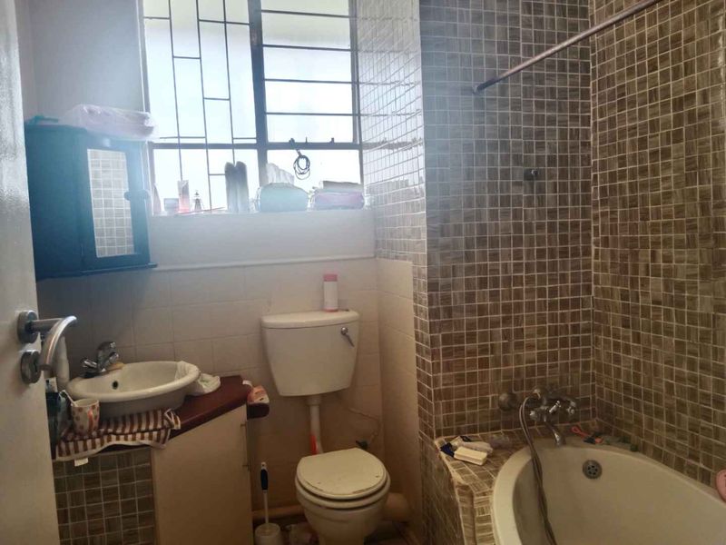 A compact bathroom featuring a built-in bathtub with mosaic tiling, a corner vanity unit, and a standard toilet, requiring cosmetic updates.