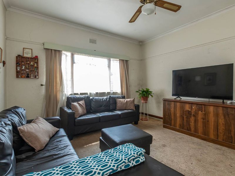 A living room featuring a ceiling fan, large window, and carpeted flooring, showing signs of wear on the walls and floor.