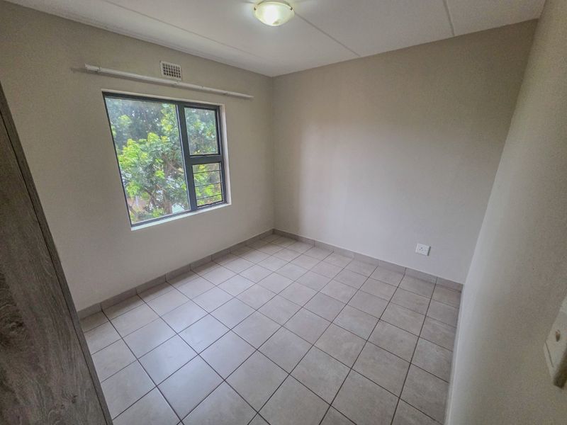 A clean, empty bedroom featuring neutral tiled flooring, an aluminium window with security bars, and a simple ceiling light fixture.