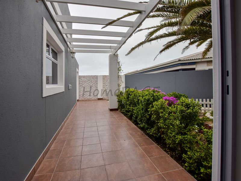 A covered outdoor patio area featuring tiled flooring and a white pergola structure, adjacent to a landscaped garden and boundary wall.