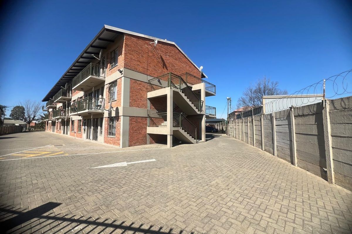 Exterior view of a multi-story brick apartment building featuring external concrete staircases, private balconies, and a paved parking area enclosed by a boundary wall.