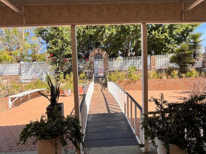 A covered patio area featuring brick paving and a pedestrian bridge leading to a gated garden entrance with brick retaining walls.
