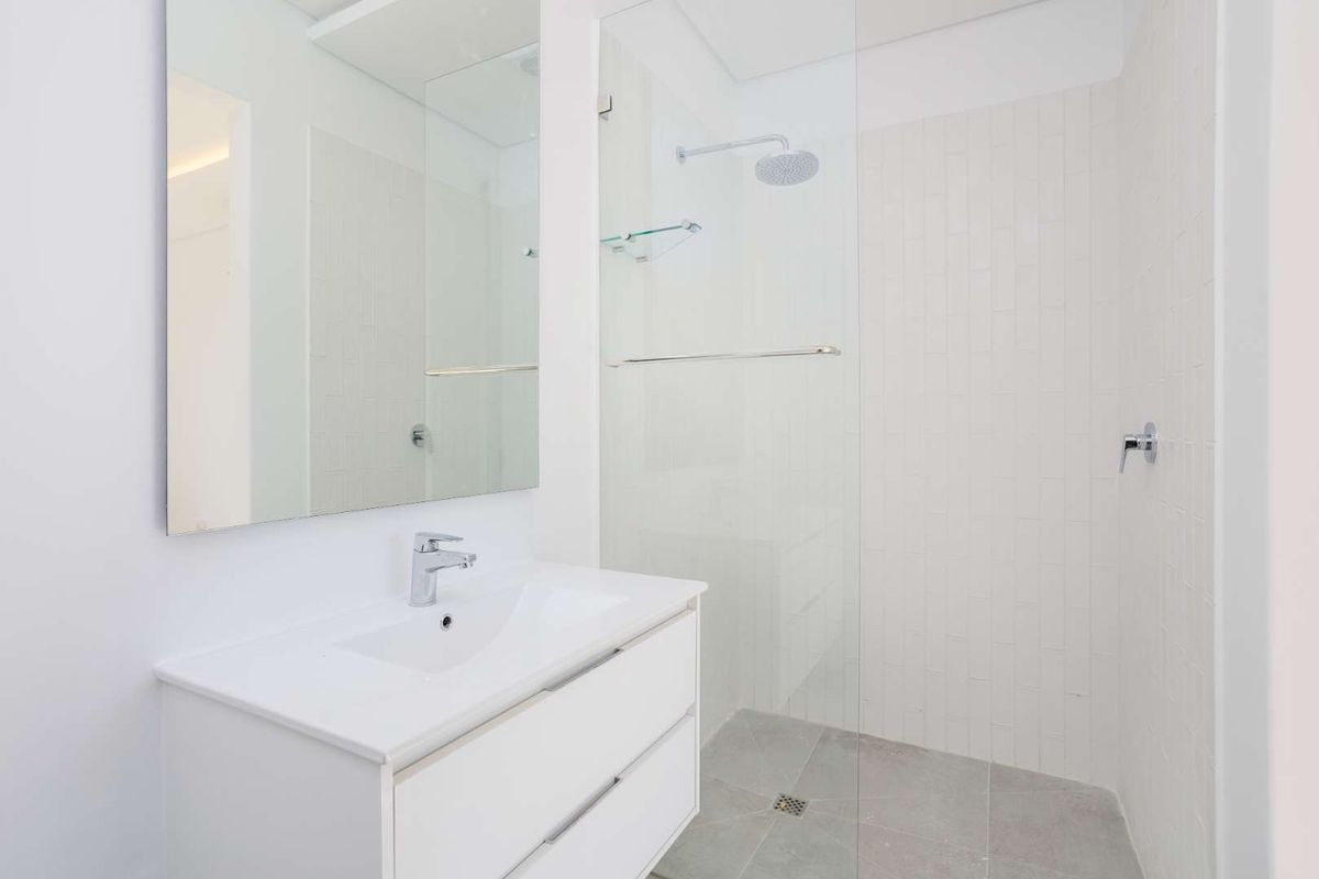 A modern, minimalist bathroom featuring a sleek white vanity, large wall mirror, and a spacious walk-in shower with a glass screen and rain shower head.