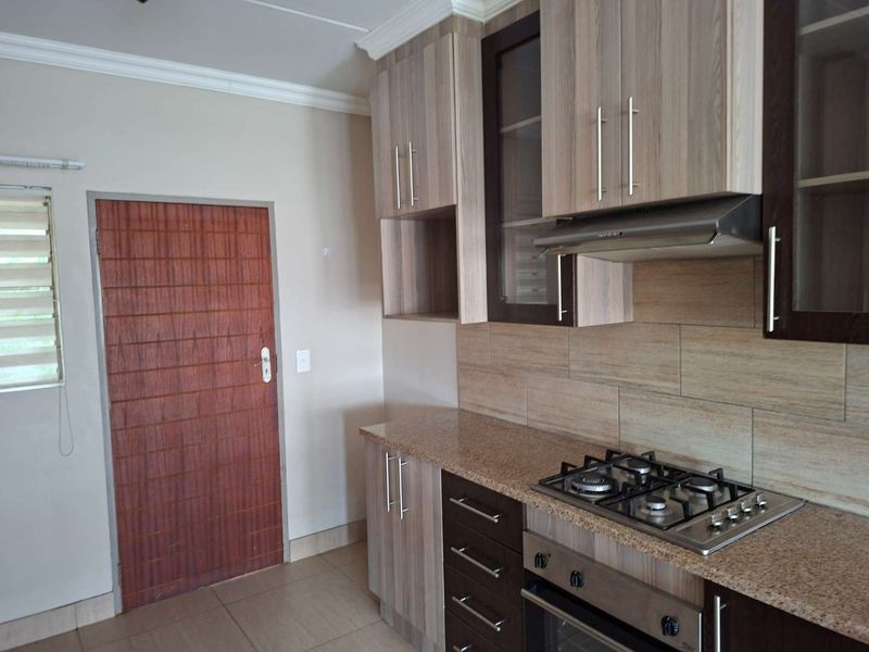 Modern kitchen featuring wood-finish cabinetry, granite countertops, a built-in gas hob, oven, and extractor fan with neutral tiled flooring.