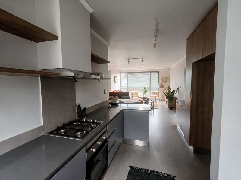 Modern open-plan kitchen featuring sleek grey cabinetry, stone countertops, integrated gas hob, and oven, flowing seamlessly into a bright living area with large windows.