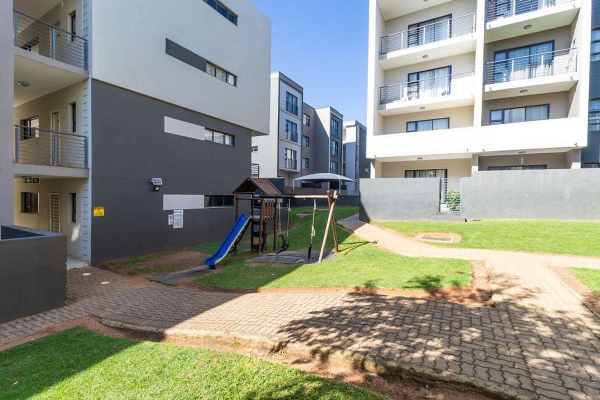 A well-maintained communal garden area between modern apartment blocks, featuring paved walkways, manicured lawns, and access to private balconies.