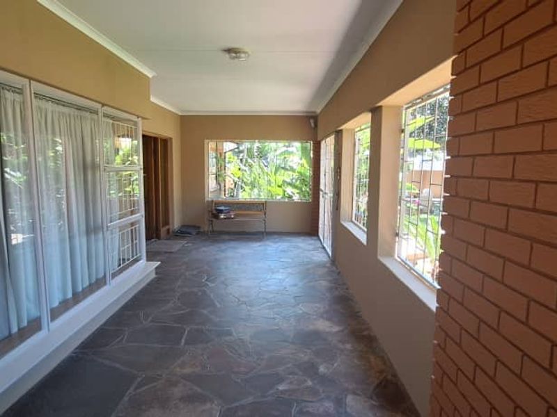 A covered patio area featuring stone flooring, a brick feature wall, and large windows with security bars, offering a bright and functional outdoor space.