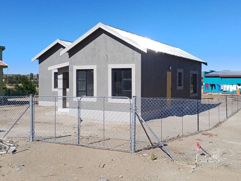 A newly constructed single-story house featuring a grey facade, metal roofing, and a perimeter chain link fence in a developing residential area.