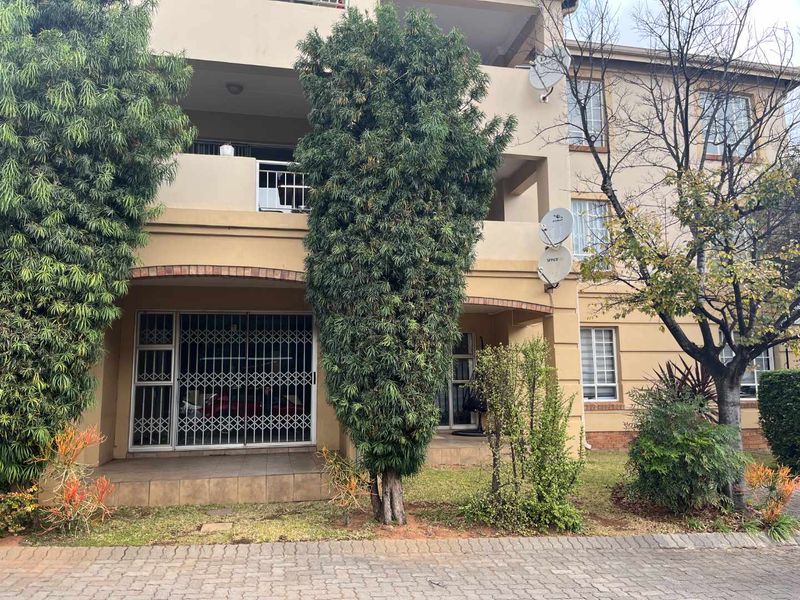 Front elevation of an apartment building featuring a ground-floor patio with security gate, upper-level balcony, and paved driveway area.