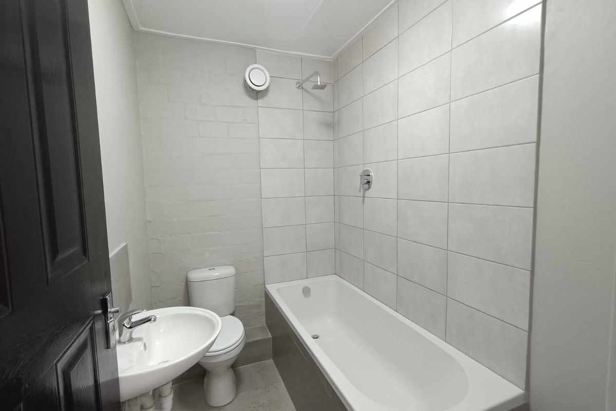 A clean, modern bathroom featuring a white bathtub with shower head, pedestal basin, and toilet, finished with neutral wall tiling.