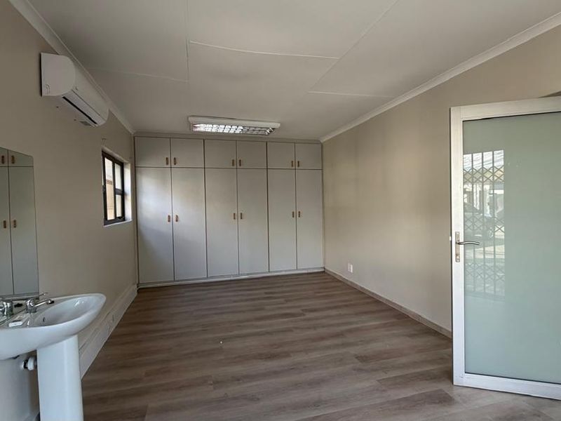 A clean, empty office space featuring laminate flooring, built-in storage cabinetry, a private pedestal sink, and a wall-mounted air conditioning unit.