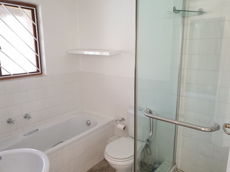 A functional bathroom featuring a bathtub, separate shower enclosure, toilet, and washbasin with white wall tiling and a window with security bars.