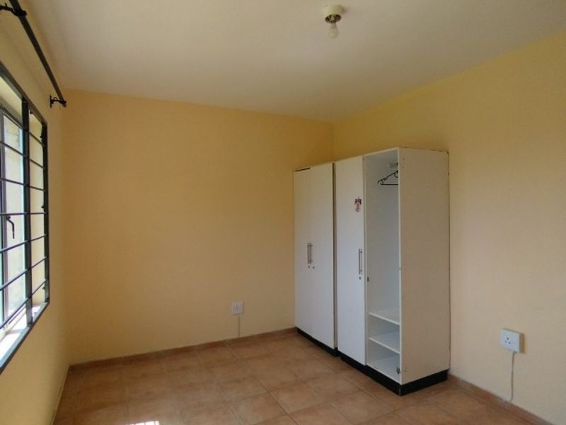 A simple, empty bedroom featuring tiled flooring, a built-in wardrobe, and a window fitted with security bars.