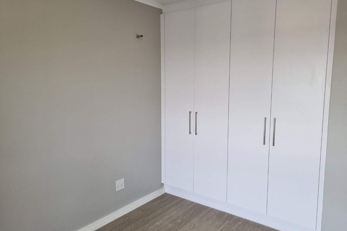 A clean, modern bedroom featuring neutral grey walls, light wood-look laminate flooring, and ample white built-in cupboard storage.