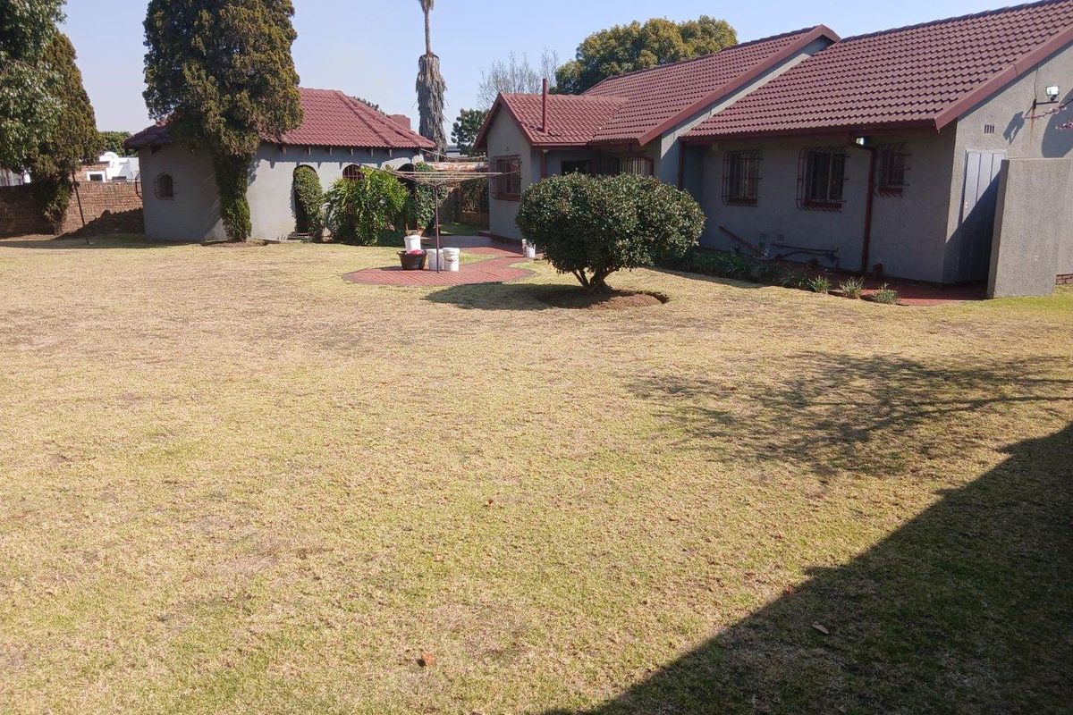 Spacious rear garden featuring a large lawn, paved patio area, and a single-story house with a tiled roof and security bars on windows.