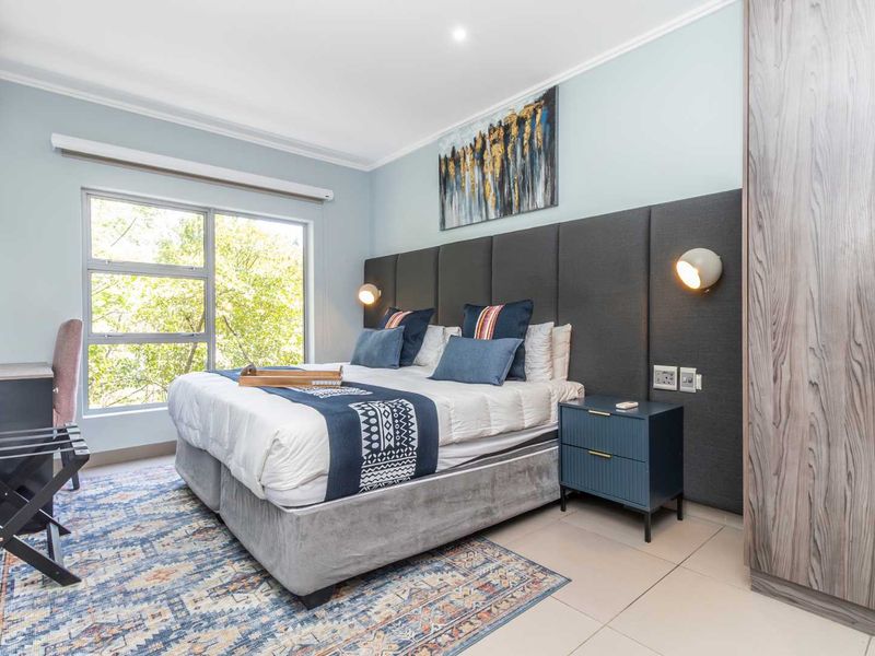 A well-maintained bedroom featuring a large built-in wardrobe, upholstered headboard, wall-mounted lighting, and tiled flooring with a pleasant view through the window.