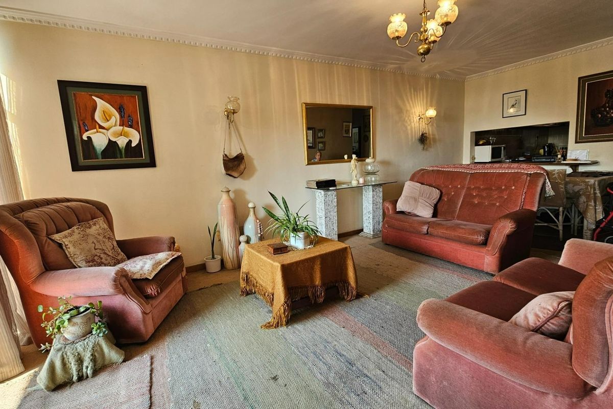 A spacious living room featuring traditional ceiling cornicing, wall-mounted lighting, and direct access to an adjacent kitchen area.